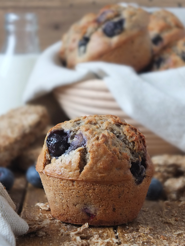 Close up photograph of a gorgeously brown weetabix muffin with pops of juicy blueberries sitting in front of a basket with the rest of the muffins in it. There are bits of crumbled Weetabix lying around in the foreground, for stylish flair (and because obviously I loved cleaning up the mess after my food photography sessions!)