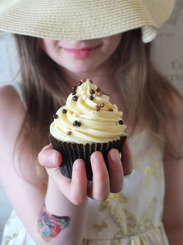 Image of a young girl in a dress and summer hat holding a chocolate cupcake in a brown paper case piled high with creamy vanilla frosting and chocolate sprinkles in front of her.