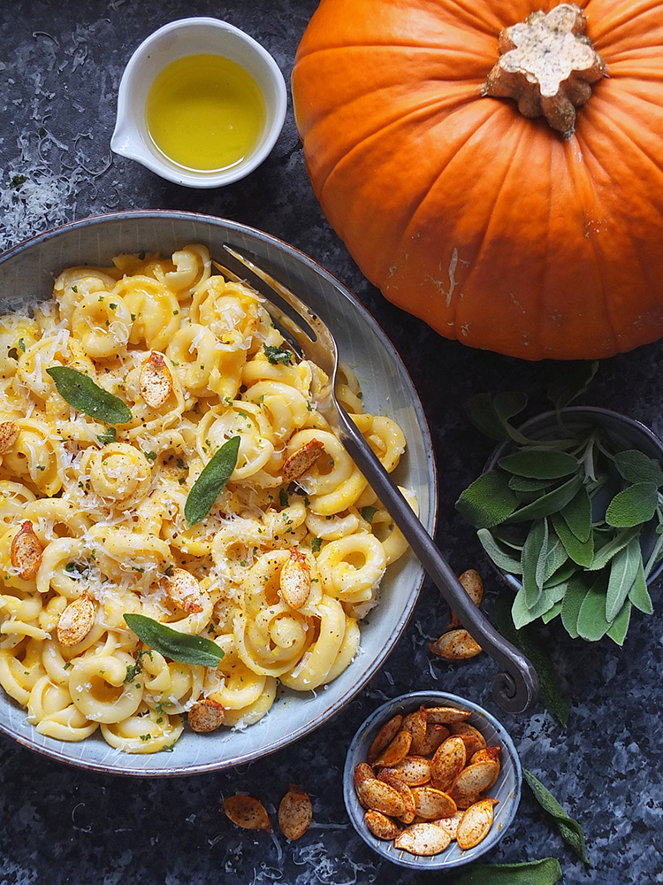 Top down image of a large bowl of Creamy Pumpkin Pasta with Fried Sage and Chilli Pumpkin Seeds. There is a whole pumpkin for decoration in the top right hand corner, a small bowl of olive oil in the top left, a bowl of fresh sage leaves and a tiny bowl with chilli pumpkin seeds overspilling onto the blue counter top.