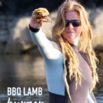 pinterest image of grilled lamb burger held in hand by red haired woman in a wetsuit