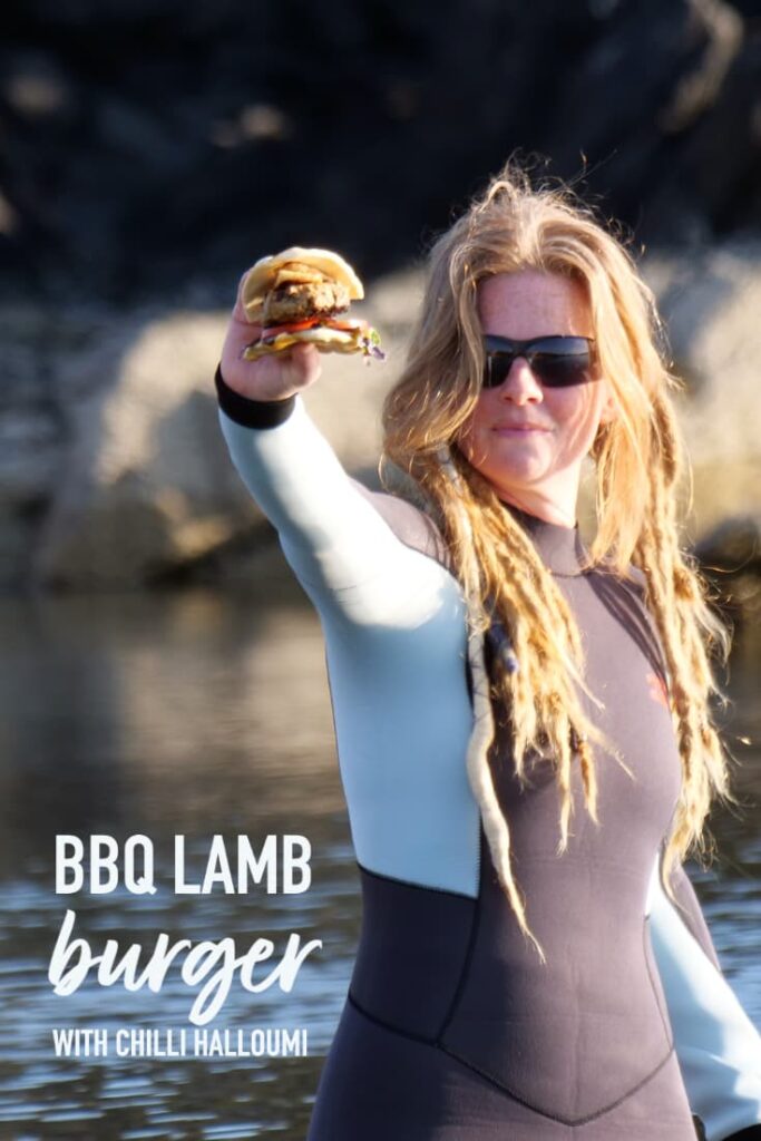 pinterest image of grilled lamb burger held in hand by red haired woman in a wetsuit