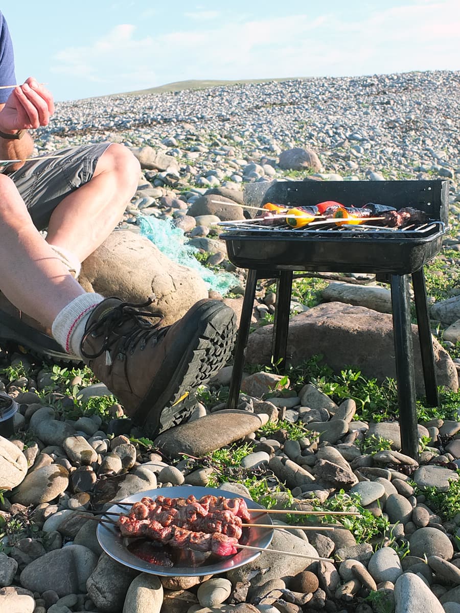 Man sitting on pebbly beach eating a recipe for lamb skewers straight off the barbecue. 