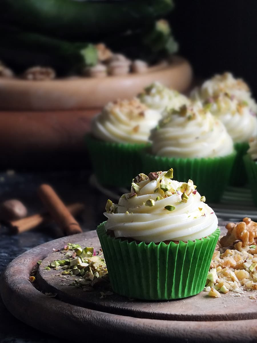 Image of spiced courgette cupcakes in green cupcake cases piled high with a swirl of cream cheese frosting and sprinkled with chopped pistachio nuts.