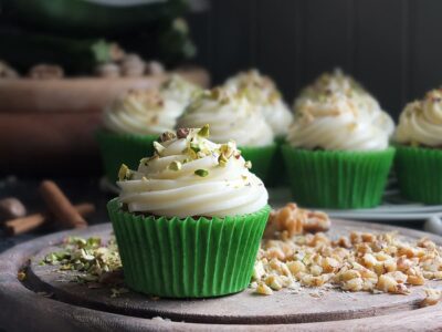 Photo of spiced zucchini cupcake with cream cheese frosting on a wooden serving board surrounded in chopped nuts.