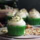 Photo of spiced zucchini cupcake with cream cheese frosting on a wooden serving board surrounded in chopped nuts.