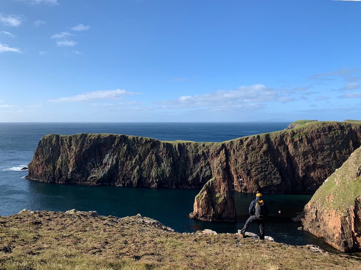 Image of man standing at the edge of a cliff looking out to some sea stacks and the open sea.