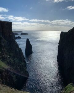 Image of the silhouette of a man standing atop a massive cliff with sea stacks in the distance.