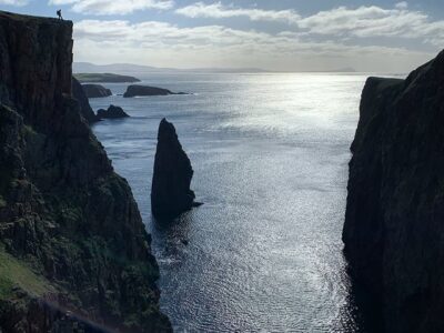 Image of the silhouette of a man standing atop a massive cliff with sea stacks in the distance.