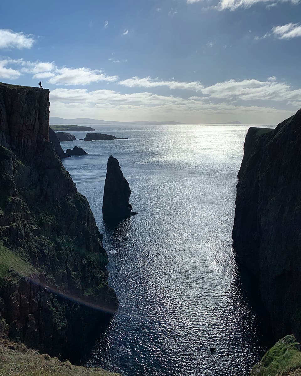 Image of the silhouette of a man standing atop a massive cliff with sea stacks in the distance. 