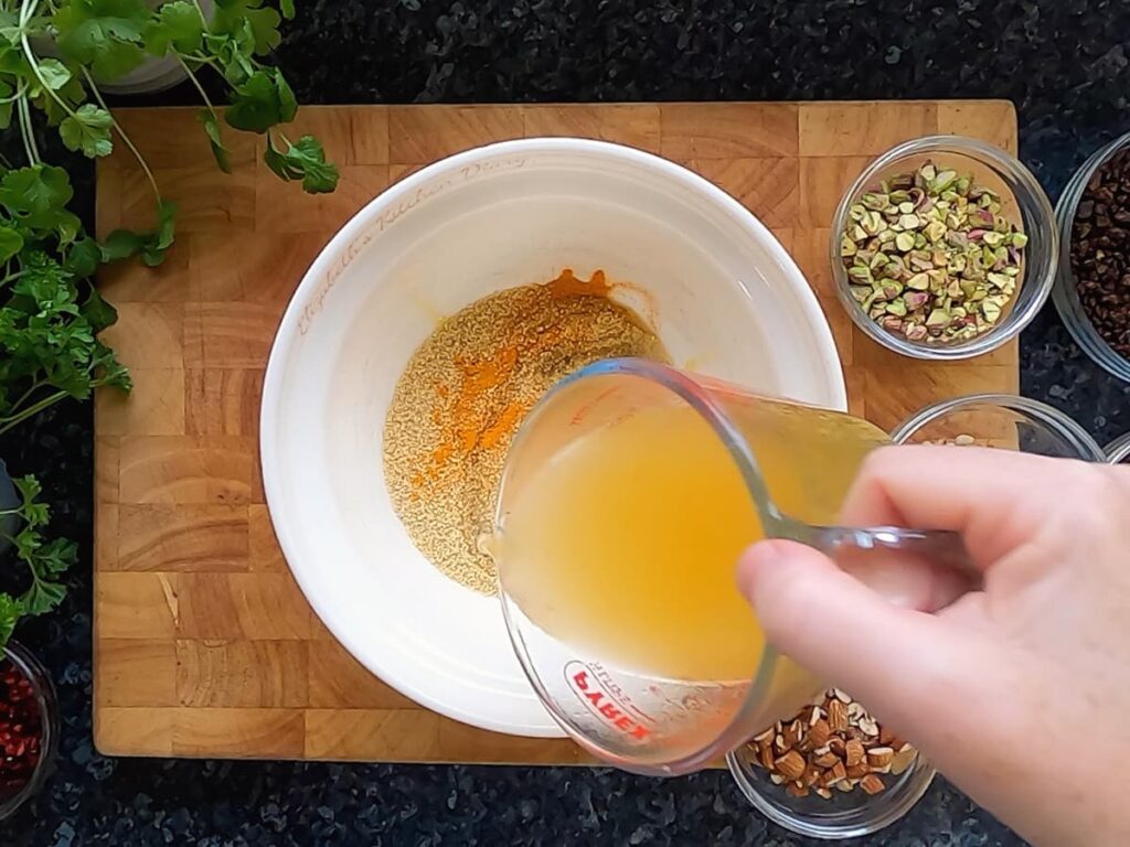 Image of vegetable stock being poured into a bowl of dry couscous with turmeric and olive oil.