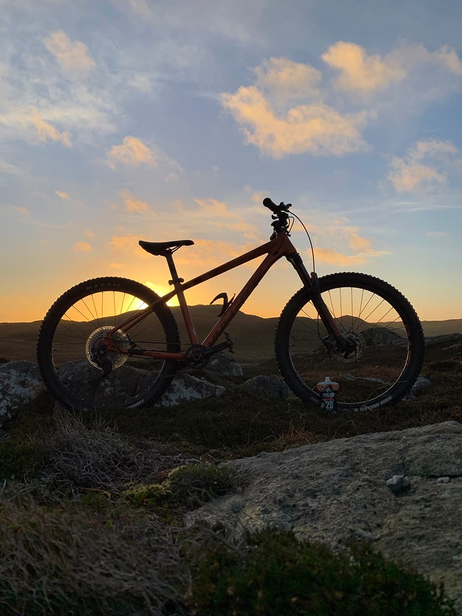 Image of a mountain bike silhouette against a sunset sky with an Arla Protein 20 pouch by the front tyre.