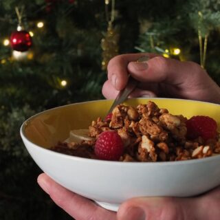 Image of man's hands holding a bowl of peanut butter granola. There's a little bit of oil on his fingers as he's a cyclist and he had a tyre puncture just before I asked him to be my hand model.