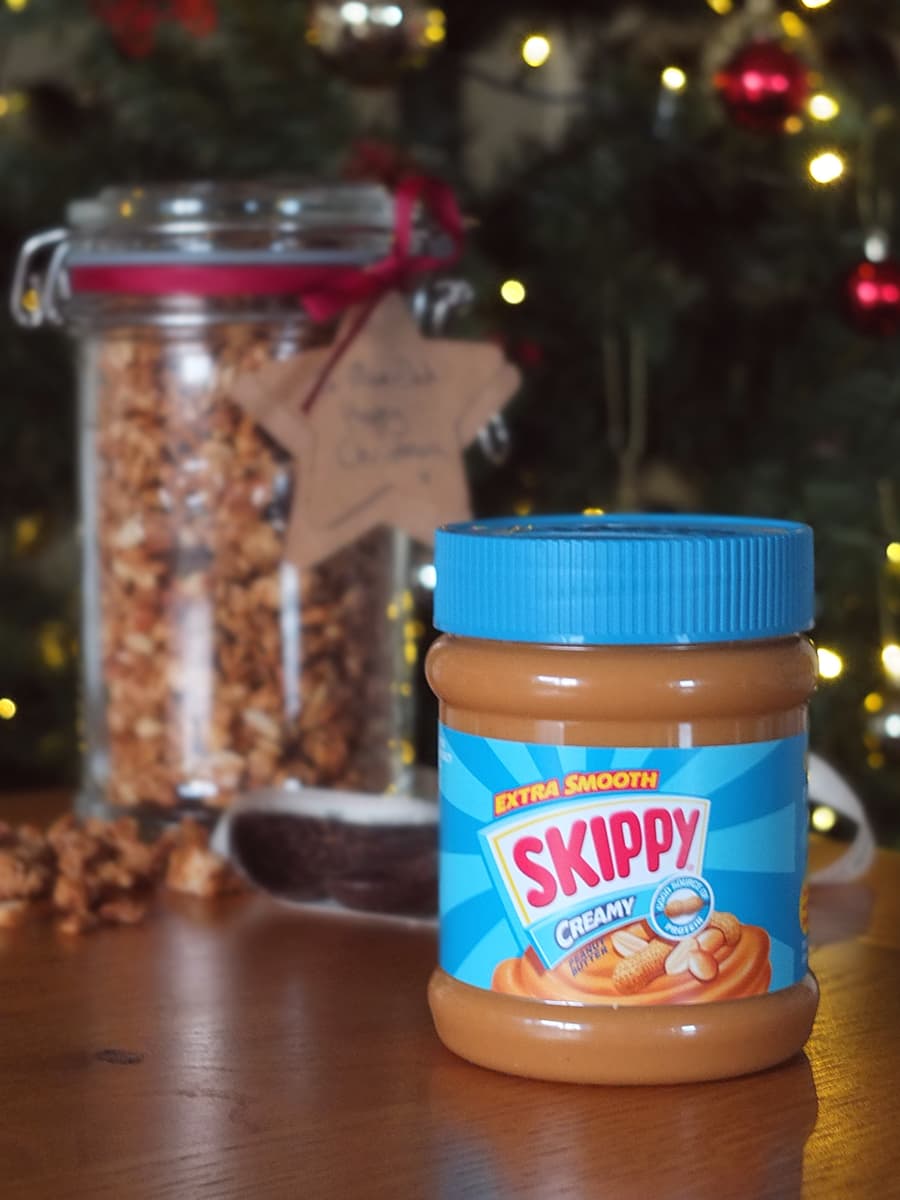 Image of a jar of SKIPPY peanut butter in the foreground with a jar of peanut butter granola in the background, in front of a Christmas tree.