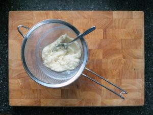 Image of celeriac puree being pushed through a fine sieve with the back of a spoon.