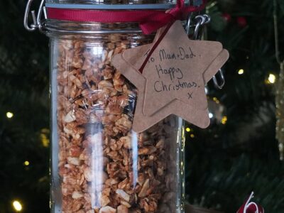 Image of large glass jar filled with peanut butter granola tied with a dark burgundy ribbon and a natural paper tag reading 'To Mum and Dad, Happy Christmas!'