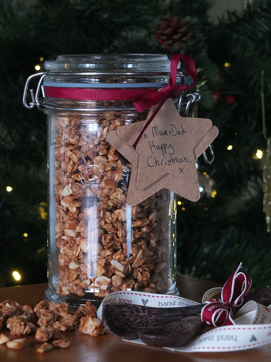 Image of large glass jar filled with peanut butter granola tied with a dark burgundy ribbon and a natural paper tag reading 'To Mum and Dad, Happy Christmas!'