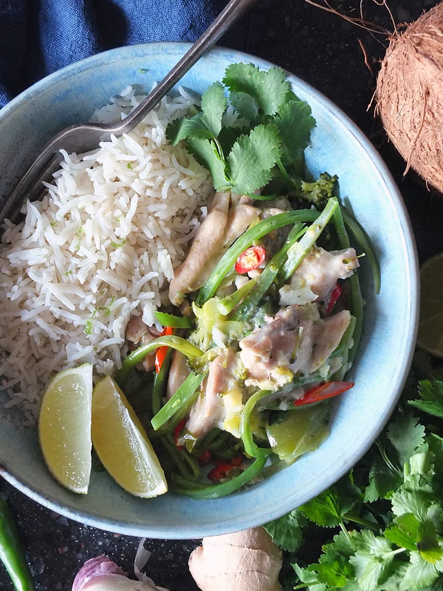 Top down image of Thai green chicken curry in a blue bowl with a side of rice, and lime wedges and coriander leaves to garnish.