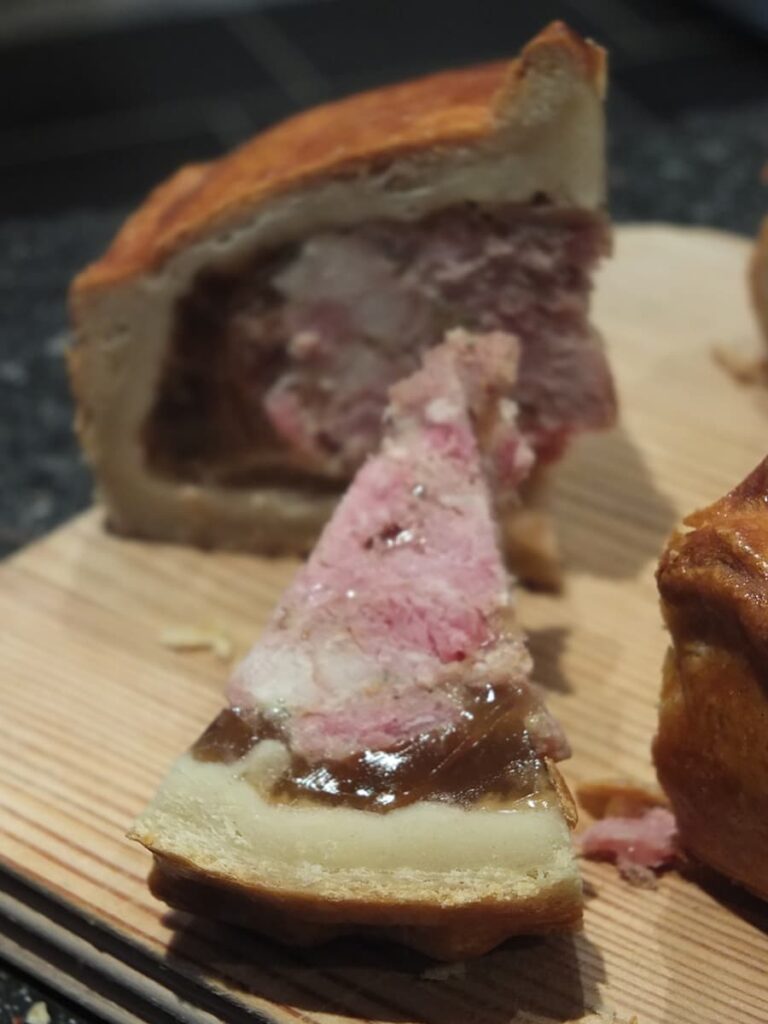 Image of a pork pie cross section showing the different layers including the hot water crust pastry outer, the different texture of the inside pastry layer, the jellied pork stock and the pork pie filling.
