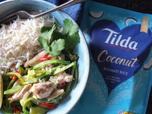 Image of Thai green curry in bowl with rice on the side. There's a vibrant blue packet of Tilda coconut rice to the right.
