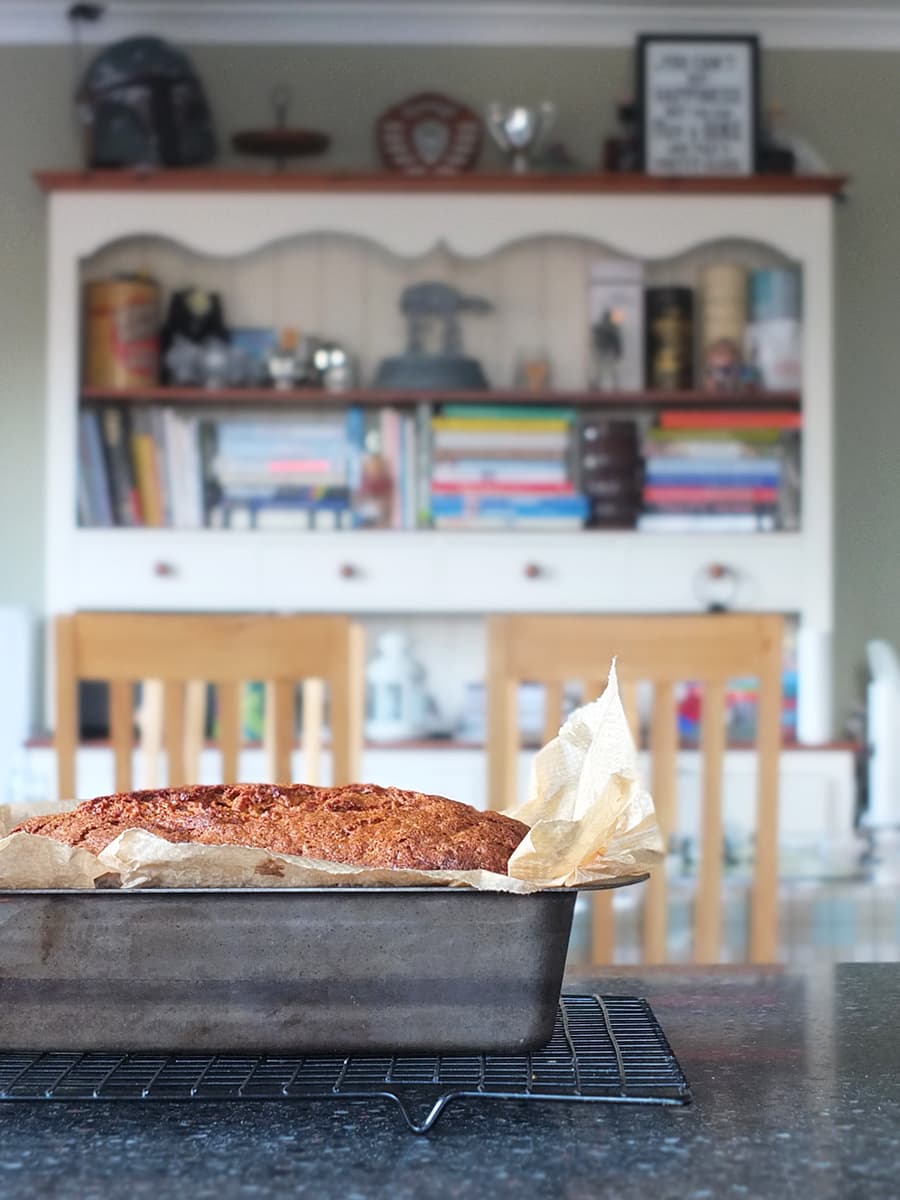 Image of banana bread in wrought iron pan with a Welsh dresser blurred in the background.