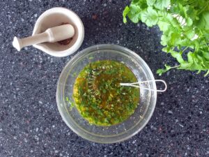 Top down image of coriander vinaigrette in a small glass bowl with a ceramic mortar and pestle to the top left hand side. There's a bunch of fresh coriander in the top right hand corner.