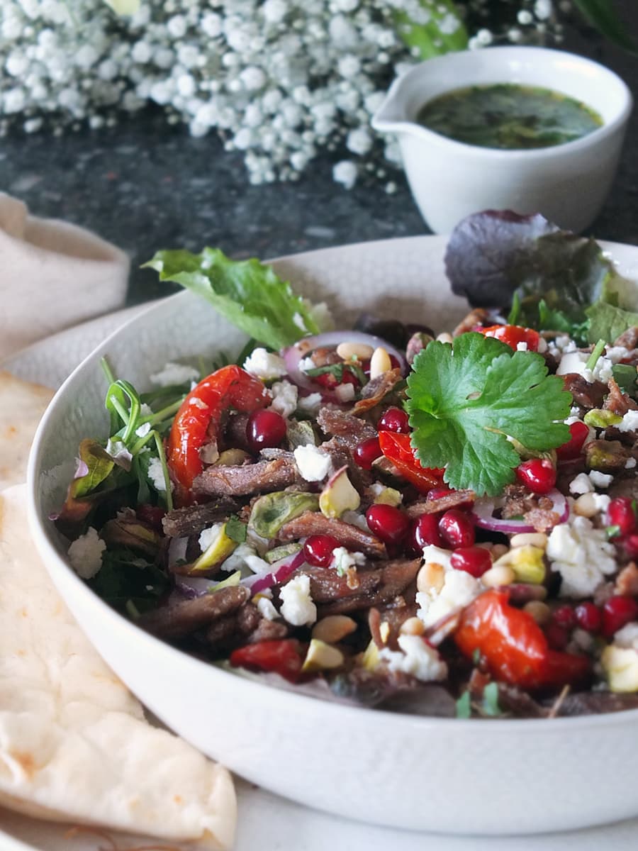 Image of a bowl full of crispy lamb salad with lots of vibrant colours and textures. There's a bouquet of flowers in the background.