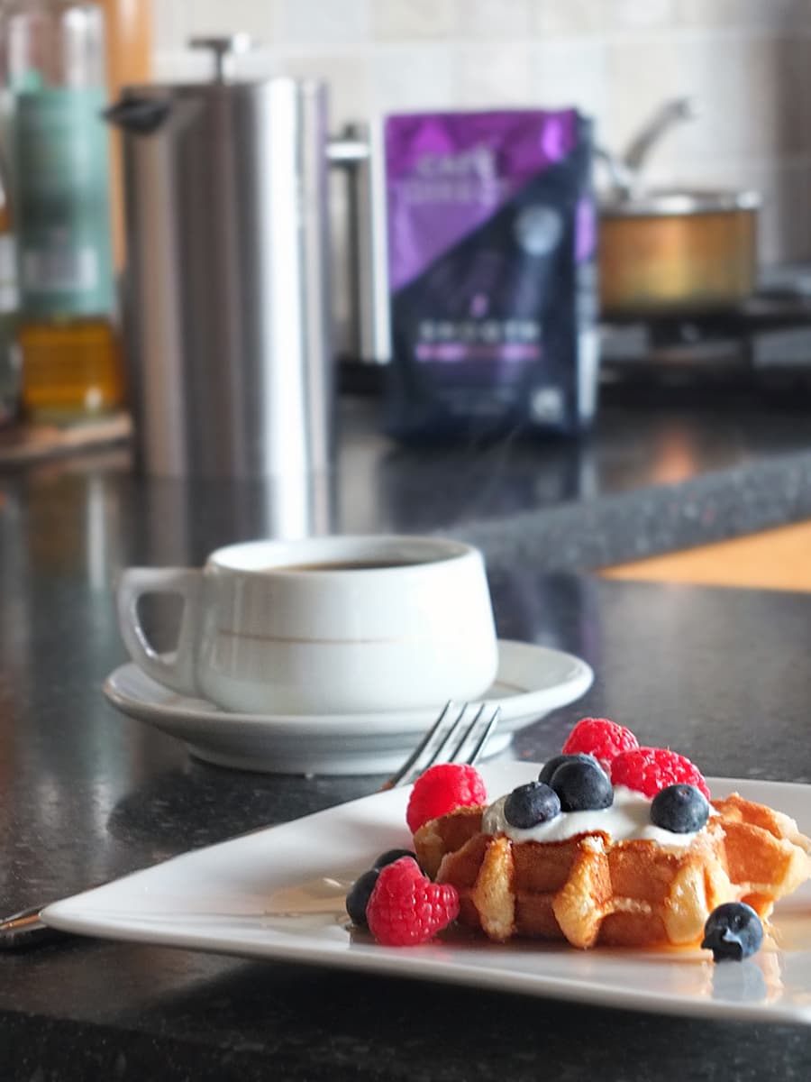 Image of sugar waffle loaded with natural yogurt and berries in the foreground with a cup of coffee, a cafetiere and the bag of Cafedirect blurred in the background.