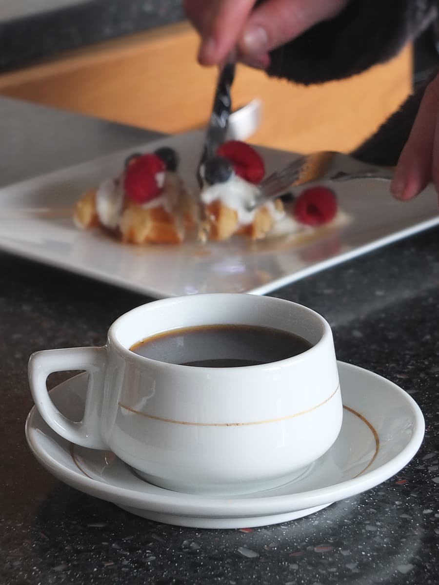 Image of cup of coffee with man eating waffle in the background.