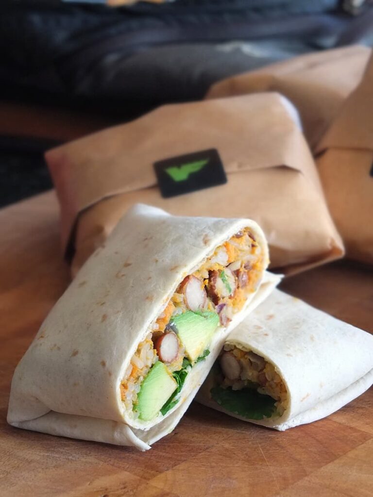 Image of a vegetarian bean burrito in the foreground, cut in half to show the fillings. Burritos wrapped in brown paper and a Veloform sticker are blurred in the background.