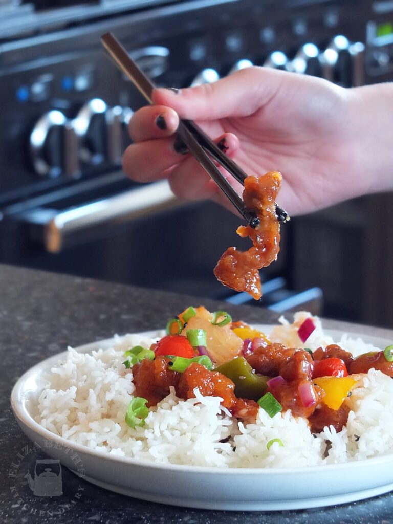 Photo of a hand holding chopsticks with a piece of crispy pork from sweet and sour pork.