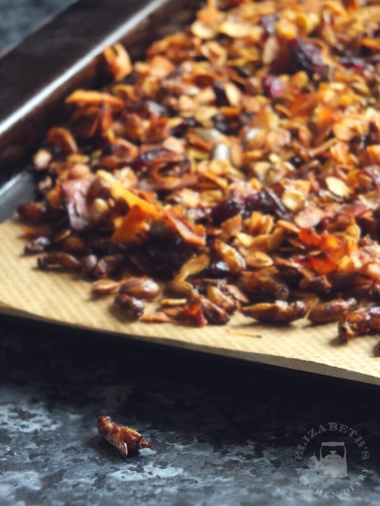 Image of a baking tray with crunchy cricket granola cooling on it. One cricket has fallen off onto the countertop and is in focus in the foreground. 