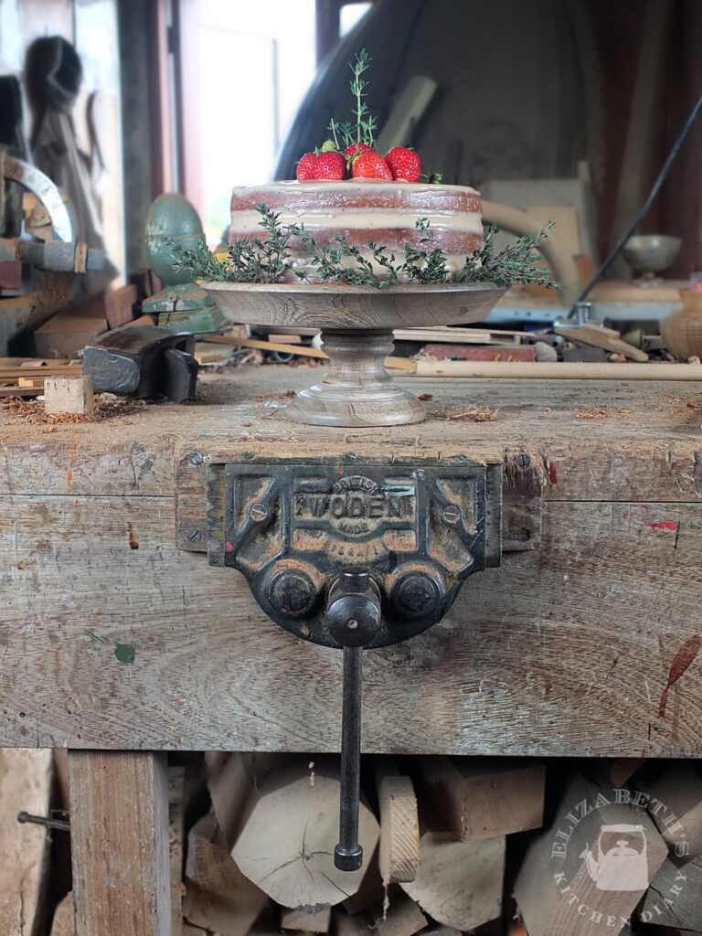 Image of a three layer eggless birthday cake on top of a wood turning bench. There is a vintage wood vice in the foreground.