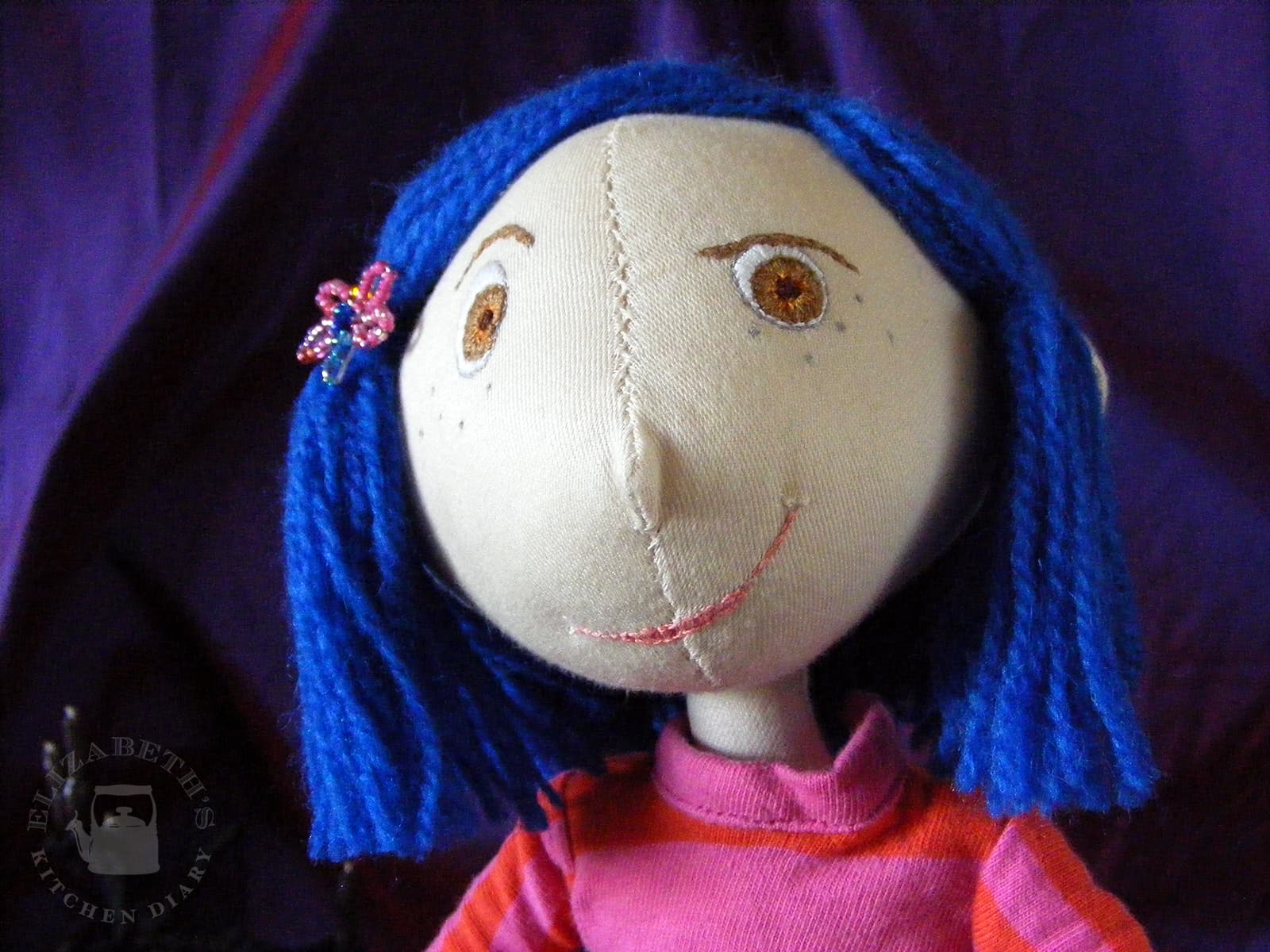 Close up image of the fabric Coraline with her embroidered face and little cheeky grin.