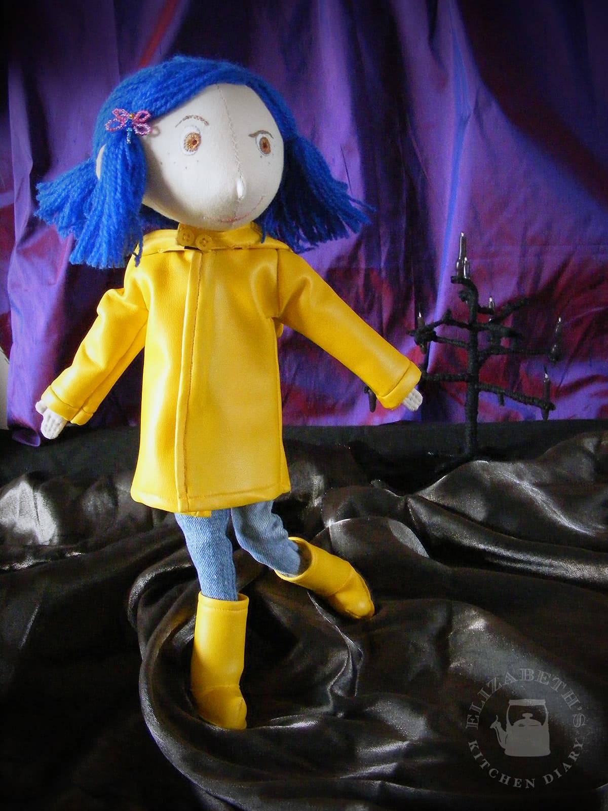 Photograph of the finished Coraline doll with yellow raincoat and boots standing up with swirled black and purple fabric around her.
