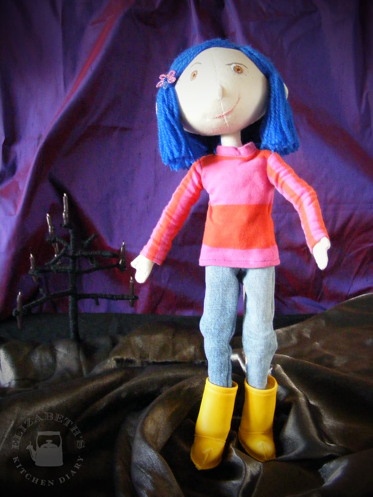 Image of a handmade Coraline doll standing up wearing a long sleeved shirt and blue jeans.