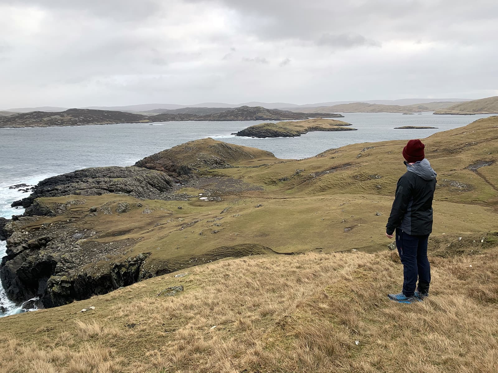 A photo of a man wearing a burgundy winter hat at the top of a hill overlooking the sea and the islands in the distance.