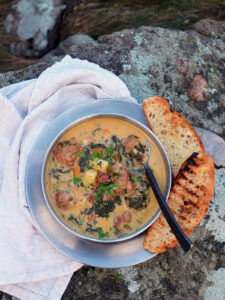 Image of a stainless steel camping bowl filled with chorizo chowder. There are a few slices of toasted focaccia on the side with gorgeous black grill lines.