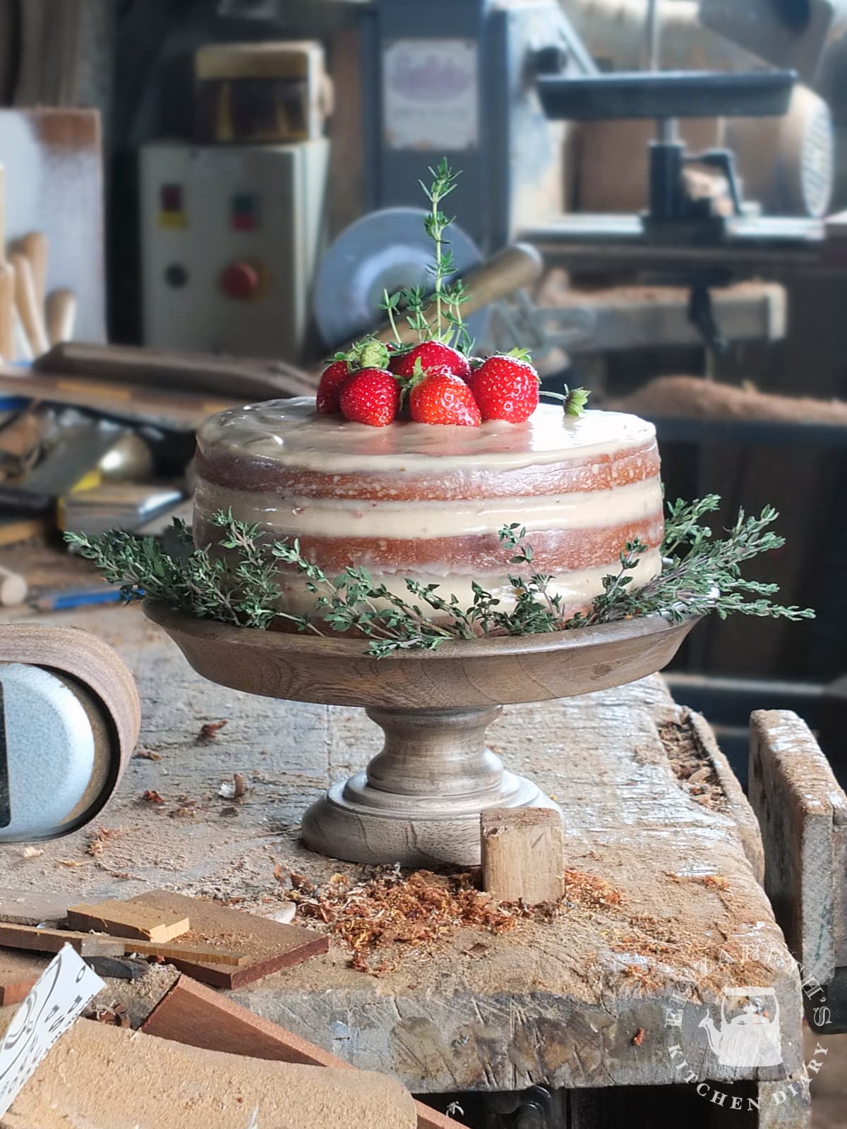 Image of a naked iced three layer birthday cake with strawberries and thyme sprigs to decorate.
