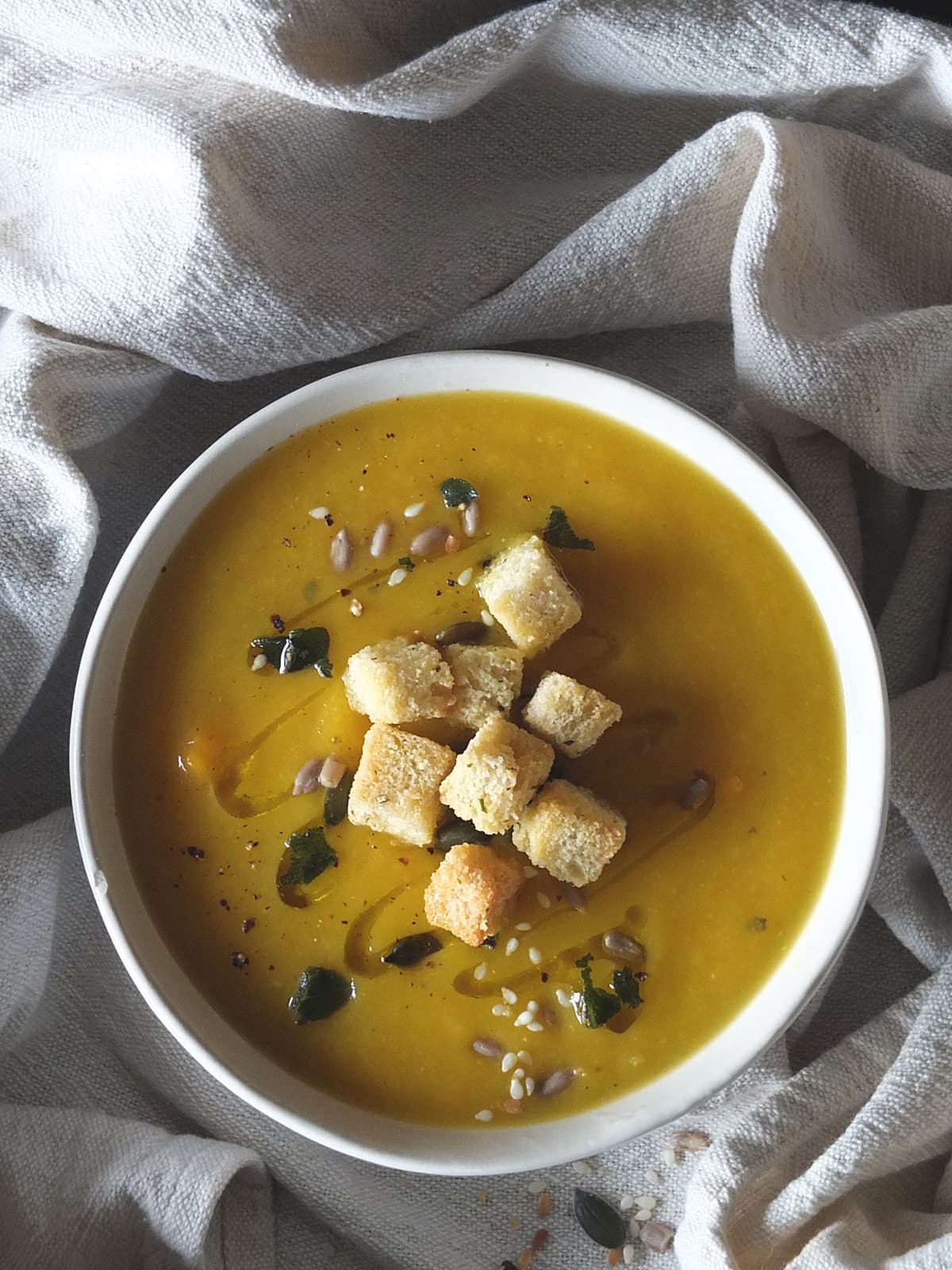 Top down photograph of a bowl of creamy pumpkin soup with fried sage and seeds topped with crunchy homemade croutons.