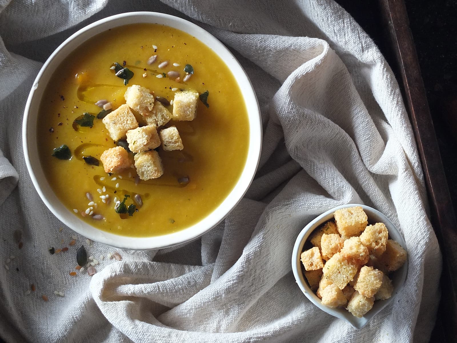 Photo of a bowl of creamy pumpkin soup topped with homemade crunchy croutons and seeds, with a little bowl of crunchy croutons.