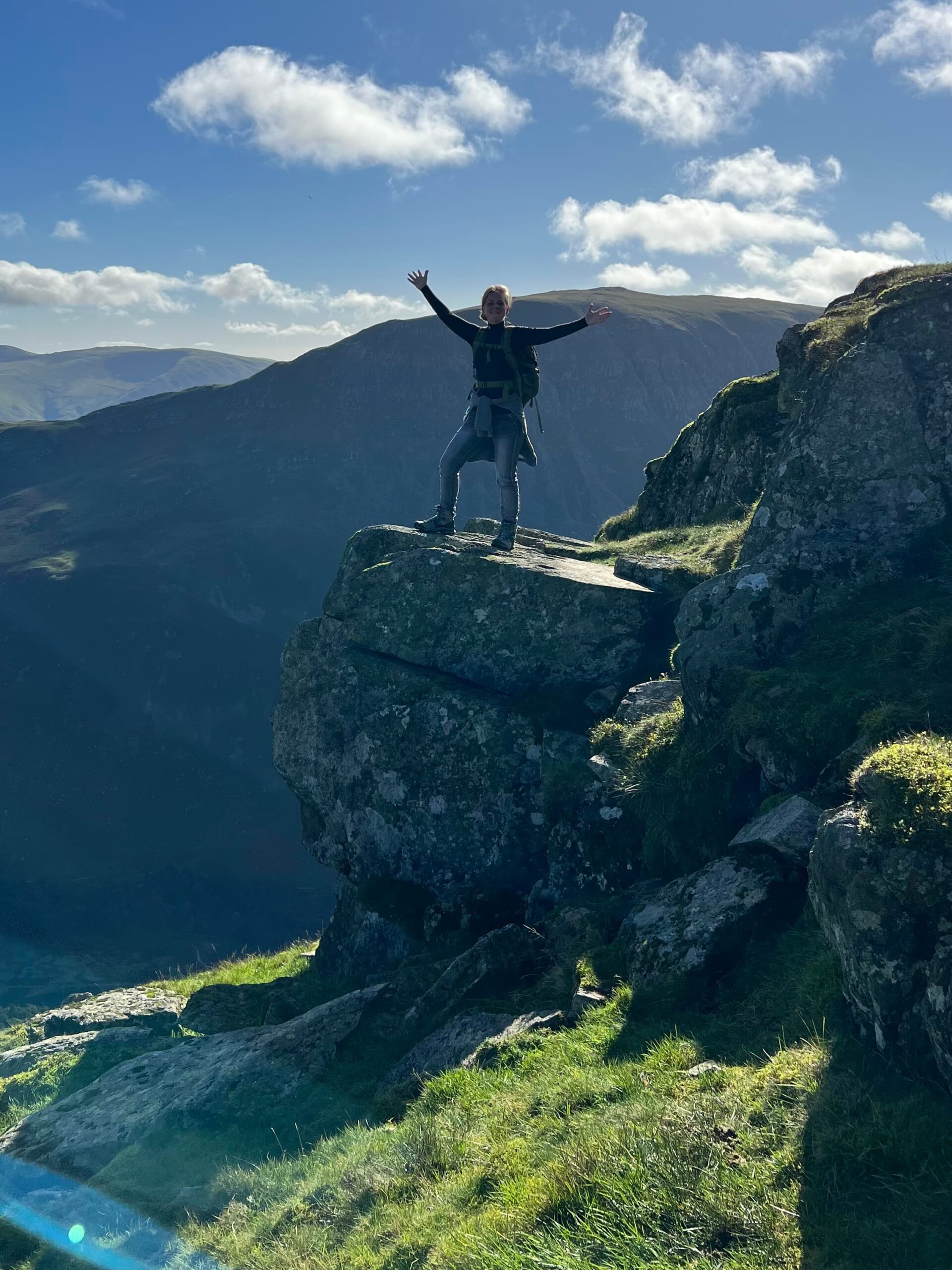 Image of a woman (me!), standing, arms outstretched on a rocky point with the valley and mountains of the Lake District in the background.