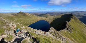 Image of Striding Edge on the way up to the summit of Helvellyn in the sunshine. Image also shows the Red Tarn and Catstye Cam with the Lake District in the distance.
