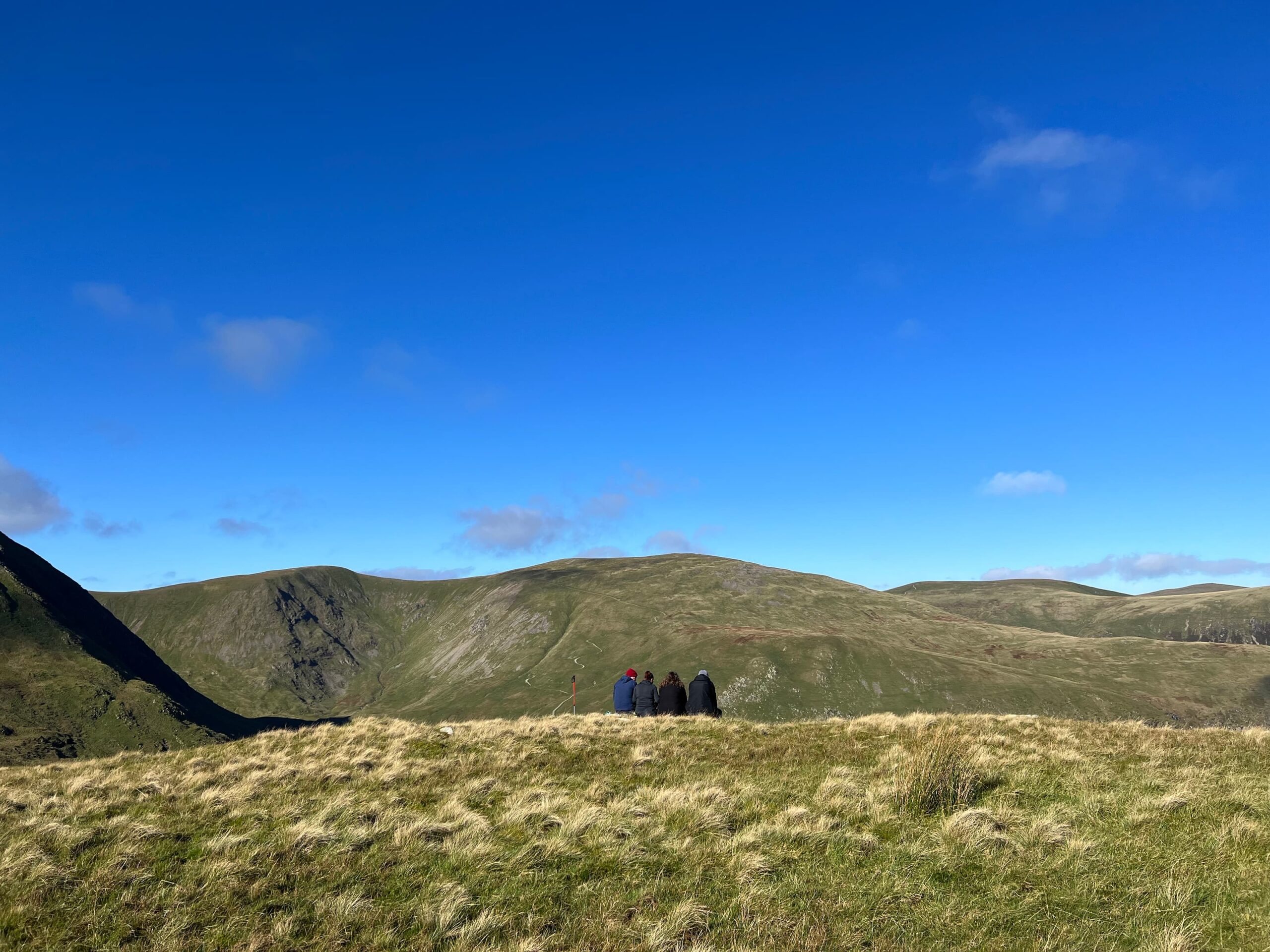 A group of four hillwalkers on Striding Edge taking a break enjoying the views. There's a walking pole to the right of them.