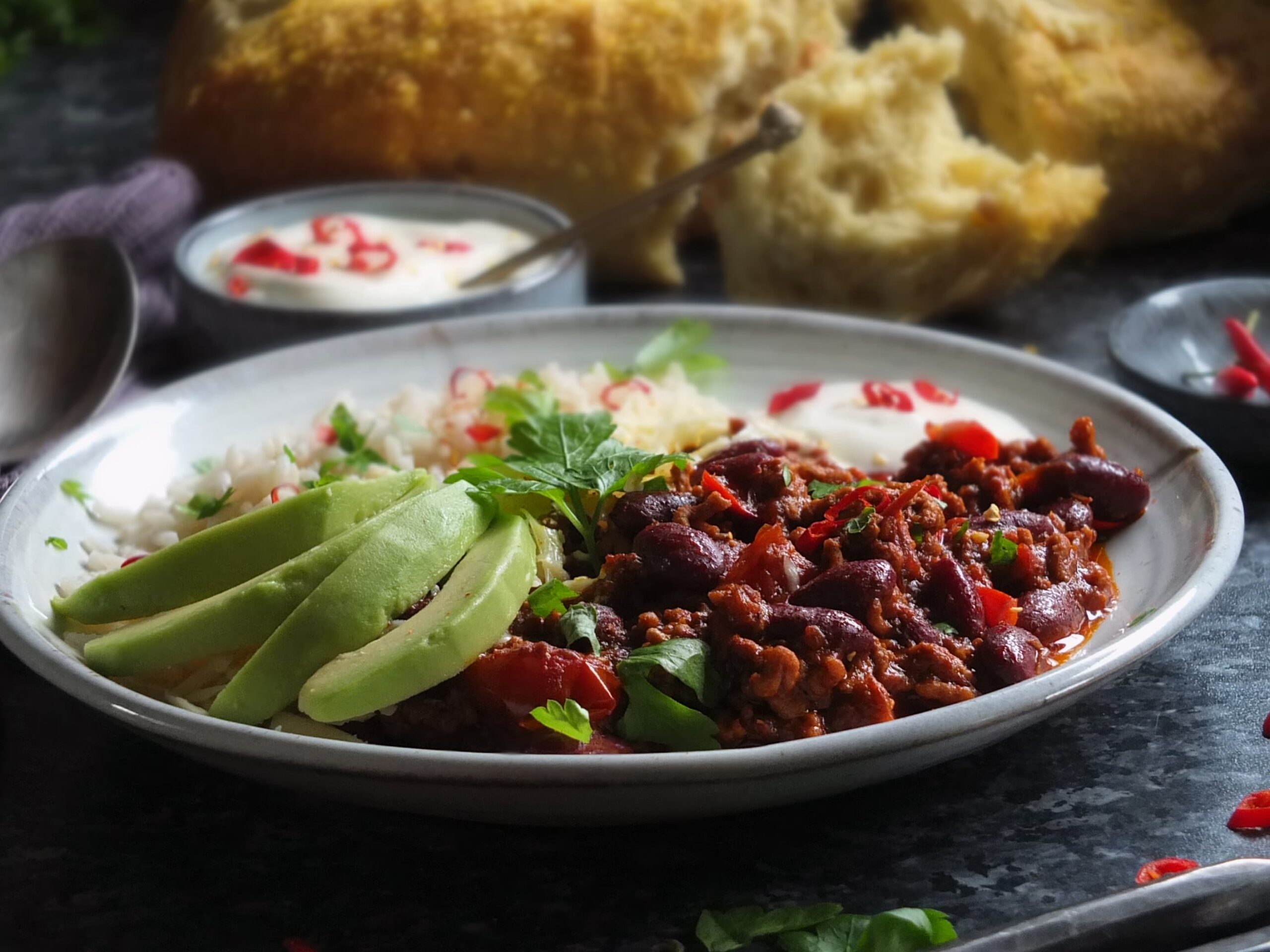 Dark moody image of chilli con carne served on a plate with rice and avocado, garnished with coriander leaves.