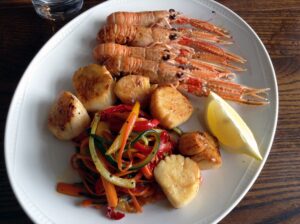 Top down image of pan-seared scallops and langoustines with stir-fried vegetables on a white plate.