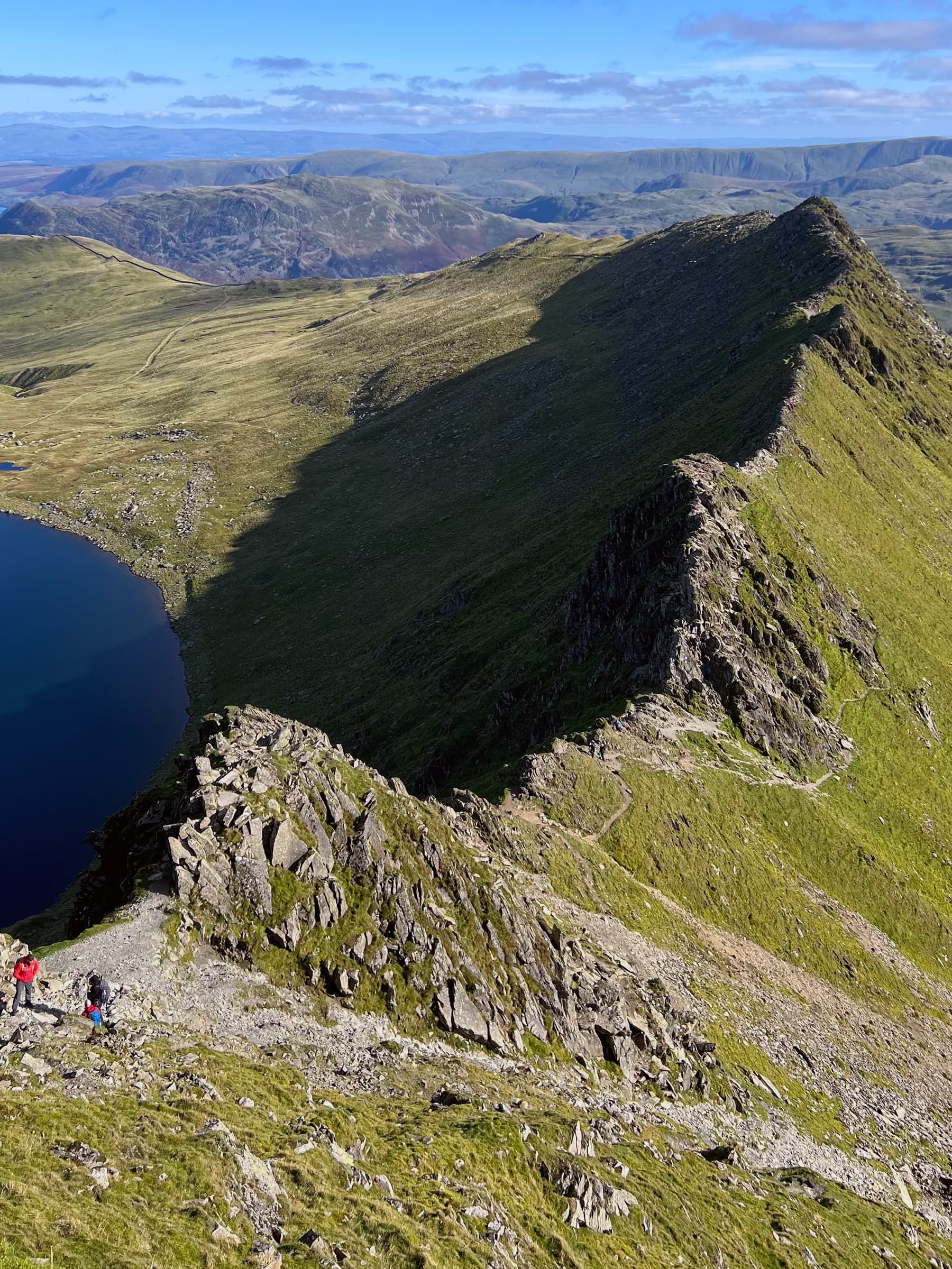 Image of Striding Edge, a knife edge of rock from halfway up the Helvellyn side. There are climbers, small, in the foreground and the Lake District mountains can be seen in the distance.