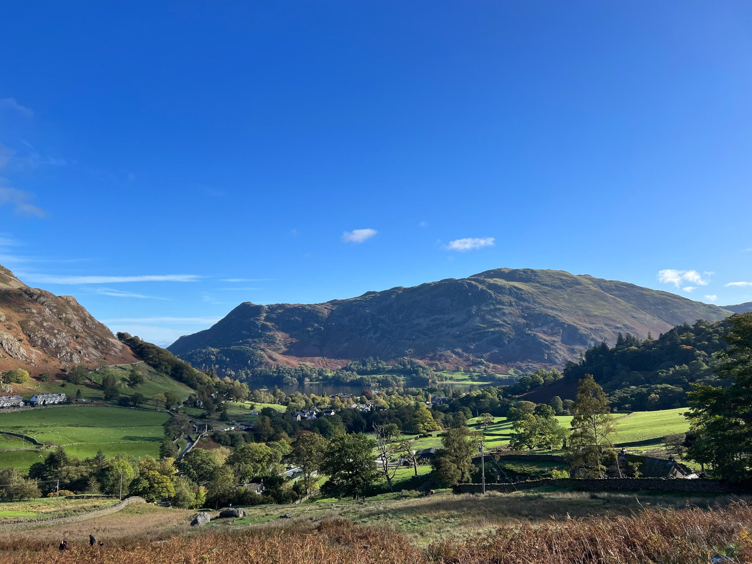 Image midway up Mires beck looking back down at the village of Glenridding. You can see Ullswater beginning to appear in the distance. The sky is blue. 