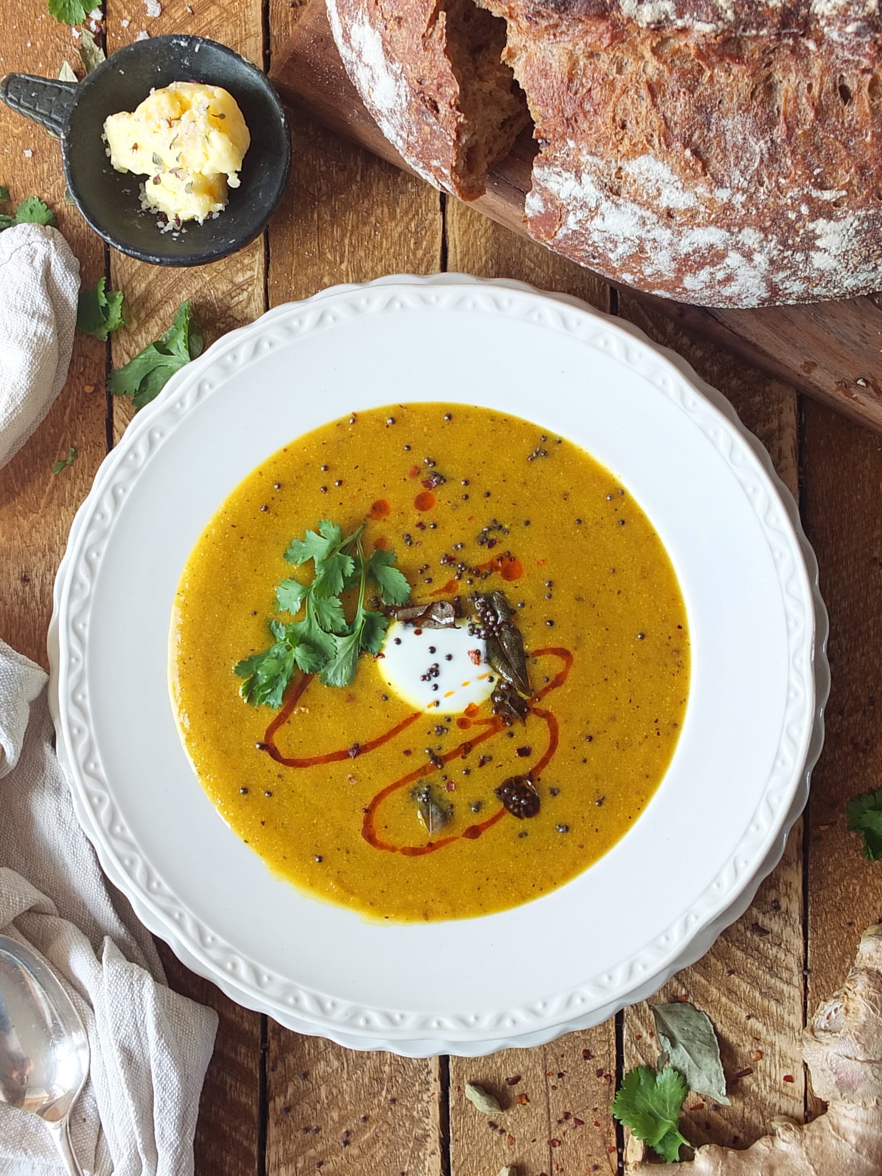 Top down image of a bowl of orange spicy carrot soup with a swirl of red chilli oil and a dollop of white yogurt. There's also a green sprig of coriander and some fried black mustard seeds and curry leaves as a garnish. It looks delicious.