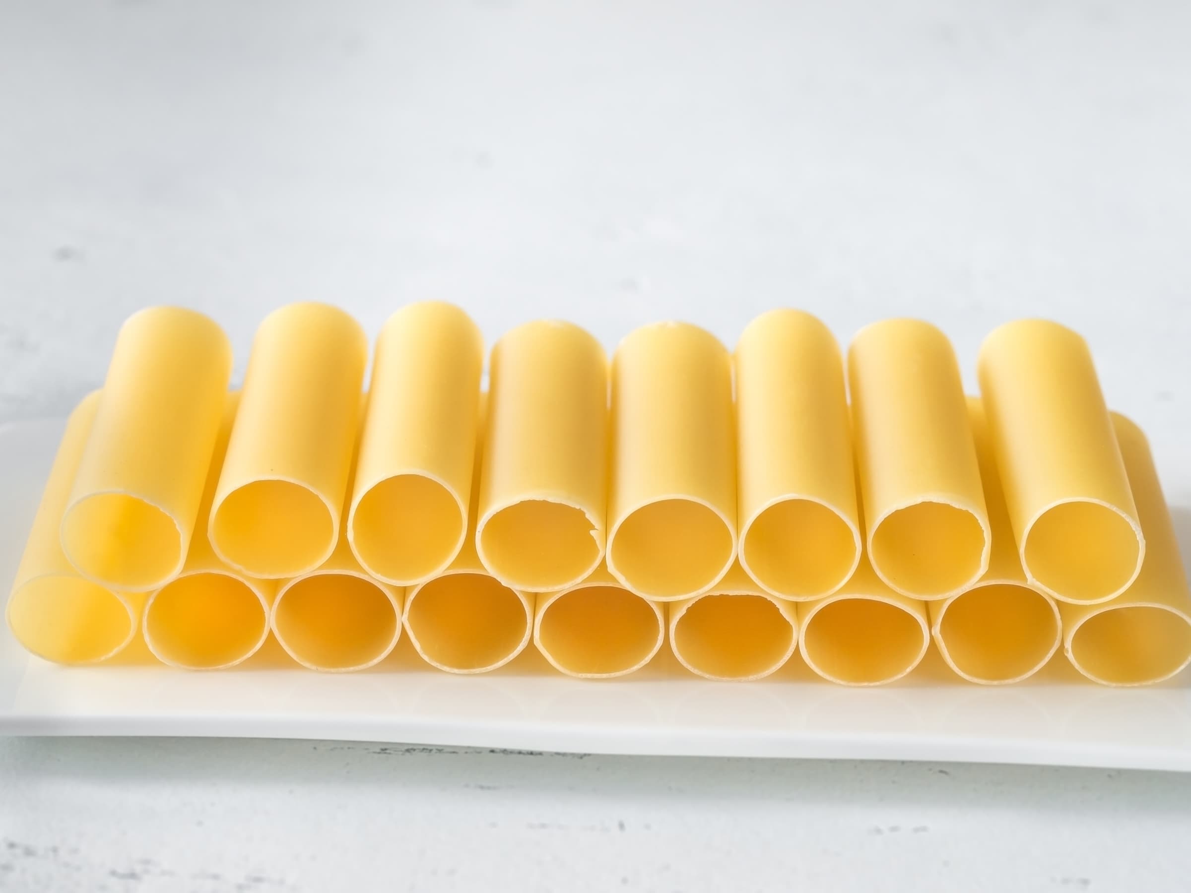 Image of 17 uncooked cannelloni tubes on a white plate with a white background.