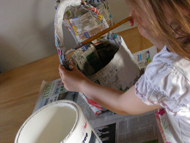 Image of child painting a papier mached woven newspaper basket with white paint.